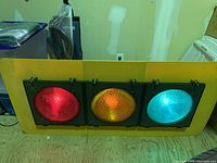 Front view showing red, amber, and green illuminated lenses in yellow metal housing on wood floor indoors.