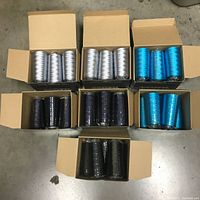 Seven open cardboard boxes containing ECOFIL embroidery thread cones in silver, black, navy, and peacock blue colors on a concrete floor.