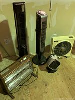 Photo showing two black tower fans, one white box fan, one silver vintage-style electric space heater, and one black Honeywell space heater