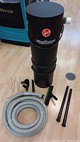 Full lot showing black Hoover Wind Tunnel central vacuum canister with gray cloth filter bag, coiled gray hose, black extension wands, floor brush, attachments, and hangers.