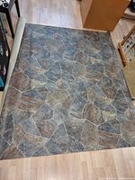 Piece of linoleum flooring unrolled on a wood floor showing stone tile pattern