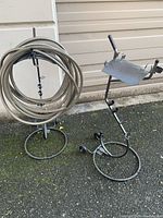 Two black iron portable hose hangers placed side by side outside against a light-colored wall; one with hose wrapped on it, the other empty showing curved support plates.