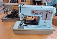 Two vintage Singer sewing machines with cases placed on a wooden table, showing the light blue sewing machine in front and the beige one behind it.