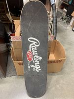 Top view of Rawlings skateboard showing black grip tape with large white Rawlings logo and red 'R' symbol, some wear visible on the edges.