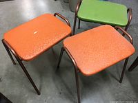 Photo of 3 mid-century stacking stools on a concrete floor; 2 stools with orange vinyl seats showing a small tear on one seat, and 1 stool with green vinyl seat.