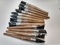 Twelve oval artist paint brushes arranged side-by-side, showing wooden handles and black bristles, some wrapped in plastic.