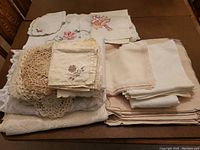 Stack of assorted white and cream linen tablecloths, placemats, and napkins with crochet and embroidered floral details, some folded and stacked on a brown surface.