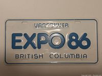Front view of EXPO 86 license plate showing raised blue lettering on white background including 'VANCOUVER', 'EXPO 86', 'BRITISH COLUMBIA'. Minor wear and surface scratches.
