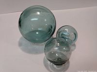 Photo showing three light blue-green glass fishing floats of varying sizes on a white background. The largest float is approximately 4.5 inches across with smaller ones beside it.