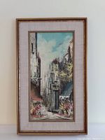 Framed oil on canvas painting showing a vertical view of a city street with buildings, flowers, and sky.