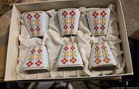 Top view of the set of six porcelain shot glasses arranged in the box with white textile lining, each decorated with floral motifs and gold rims.