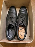 Top view of pair of black men's leather dress shoes with shoe tree inserted in right shoe inside a cardboard box.