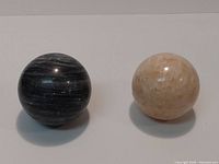 Pair of two stone spheres on white background. One is black onyx with white banding, the other is light beige calcite with natural patterns.