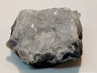 Close-up photo of a quartz stone cluster approximately 3.25 inches wide. Clear quartz crystals densely cover the top surface with a banded agate base visible around the edges.