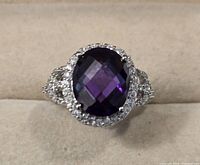 Front view of the sterling silver ring with a large oval simulated purple gemstone center and small simulated white diamonds around the halo and band.