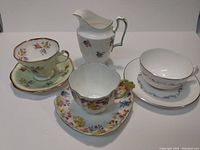 Overview of three teacups with matching saucers and a white creamer with floral decals, displayed against white background.