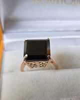 Close-up of the front profile of the ring displaying the large black square stone held by 4 prongs in rose gold plated silver setting with scrollwork filigree below the stone.