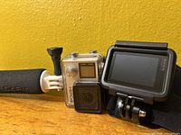 GoPro Hero4 and Hero7 Black side by side on wooden surface against yellow wall, showing front of Hero4 in housing and back of Hero7 in mount, with foam grip visible.
