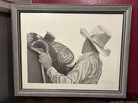 Framed black and white pencil/charcoal print showing a cowboy adjusting a saddle on a horse, highlighting detailed textures and shading.