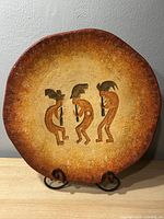 Front view of large round pottery wall plate with Kokopelli design showing three Kokopelli figures against a textured brown and beige background.