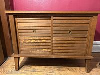 Front view of mid century record cabinet with closed doors and drawer, showing brass knobs and wooden slatted drawer fronts.