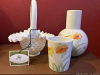 Three vintage glassware items: Fenton hobnail milk glass basket with crimped edges and curved handle, plus a hand painted bedside decanter and matching tumbler decorated with spring flowers.