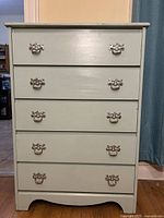 Front view of light blue wooden dresser with five drawers and ornate metal handles, showing overall condition.