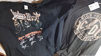 Two black rock band T-shirts, one AC/DC with High Voltage round logo and one Judas Priest 50th anniversary band photo with autographs print.