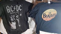 Two black T-shirts laid flat. Left is AC/DC Highway to Hell featuring a band photo and text, right is The Beatles with a circular retro logo graphic.