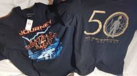 Two black vintage rock band T-shirts laid side by side. One Journey T-shirt with text and graphic on front, one Devo T-shirt with large gold metallic 50 years print on front.