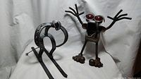 Two custom made metal art statues displayed on a white backdrop. One is a skiing cowboy made from horseshoes. The other is a quirky creature with red glass eyes and exaggerated limbs.