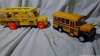 Two vintage school bus toys shown side by side on white backdrop. The Fisher-Price bus is yellow wood and plastic with red wheels and pull string. The Buddy L bus is a yellow metal collector's piece with detailed features and black tires.