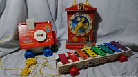 Red Goldilocks and the 3 Bears Playhouse pull toy, Fisher-Price teaching clock with yellow hands and music box, and colorful xylophone on wheels with mallets.