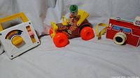 Photo showing three vintage Fisher Price toys: a white and yellow music carry radio with a rainbow decal, an orange and yellow jalopy clown car with red wheels and a clown figure, and a red plastic camera with a yellow shutter button.