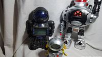 Three vintage robot toys: black coin bank robot with digital screen, small silver walking robot with digital face, and tall silver walking talking disc-shooting robot