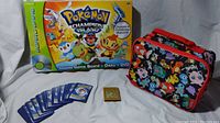 Photo of the entire set showing the Pokemon Champion Island board game box, 50 blue-backed Pokemon cards spread in a fan, the gold cartridge, and the Pokemon-themed Thermos lunch box.