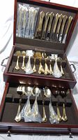 Overall view of opened wooden box displaying multiple compartments of gold tone plated forks, knives, spoons, and serving utensils, some still in plastic packaging.