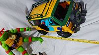 Green and yellow TMNT van with some damage, large TMNT character doll lying next to it, ruler showing size approximately 13 inches
