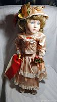 Full-body photo of porcelain doll wearing peach Victorian dress with lace and holding red box and floral bouquet.