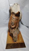 Front and left side view of the driftwood face carving, showing the carved facial features and natural wood texture on a wooden base.