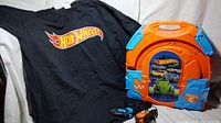 Black Hot Wheels t-shirt with large Hot Wheels logo in front next to an orange and blue plastic Hot Wheels slot cars track carry case with attached controllers.