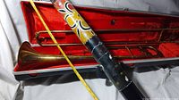 Painted didgeridoo held above the trombone case, showing detailed painted designs and measurement with a yellow tape measure, about 4 feet long. Cracked wood visible.