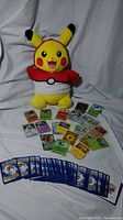 Full view of Pikachu plush doll wearing a red and white hoodie with scattered holographic and regular Pokemon cards laid in front, showing backs and fronts.