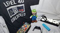 Image showing entire lot with all items arranged on white backdrop: the black Level 40 Unlocked t-shirt, Luigi plush doll, colorful Super Mario Odyssey case, Xbox logo light, PlayStation DualSense controller, and blue Wii remote.