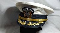Front and left side view of vintage Bancroft military officer's hat showing white top, black band, gold embroidered leaves on visor, and metal eagle insignia with anchors.