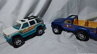Photo showing white and teal Chevy Tahoe 1500 SUV beside blue Nylint bass runner 4x4 pickup truck on white fabric background