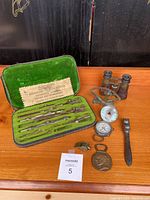 Photo showing the entire lot including Dietzgen drawing instruments set, vintage binoculars, compass, whistle, medallion, and fork all displayed on a wooden surface.