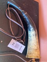 Antique curved powder horn with leather carrying strap lying on a dark surface, showing natural horn coloration and wooden stopper.