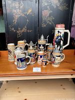 Photo showing all 12 beer steins grouped on a table with a black decorative divider behind. Various sizes and styles visible, some with lids.