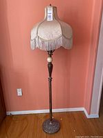 Full view of the vintage floor lamp with cream fabric lampshade featuring white fringe, wooden stand, and decorative metal base.